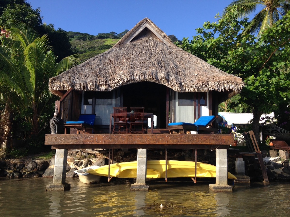 A wooden bungalow with a thatched roof is positioned on stilts above the water. An inviting deck holds two blue lounge chairs and a wooden dining set. Lush greenery surrounds the structure, contributing to a tropical atmosphere.