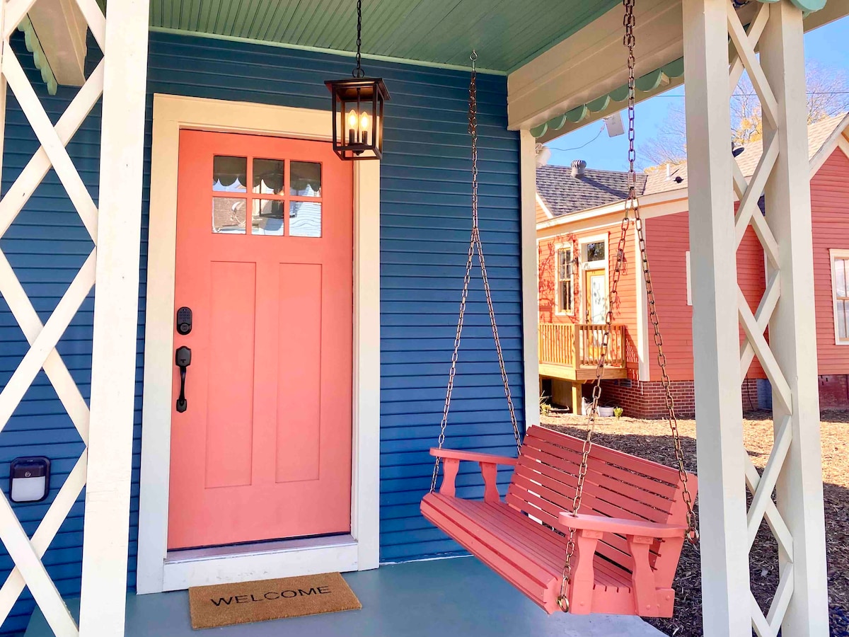 A welcoming front porch features a coral door with a glass panel, and a matching swing hangs from a decorative ceiling. A rustic light fixture provides illumination, while a doormat with 'WELCOME' offers a friendly entrance to the home.