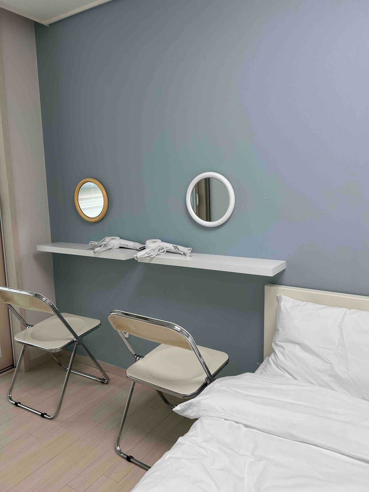 A well-organized room features two mirrored vanities mounted on a light-colored wall shelf. Two foldable chairs are positioned in front of the mirrors. A spacious bed with rumpled white linens occupies one side of the room, while soft natural light filters through nearby curtains.