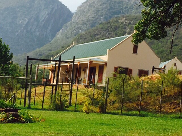A historic stone house is set against a backdrop of mountains, surrounded by lush greenery and a spacious lawn. The structure features a green roof and multiple windows. A fenced area is visible in the foreground, providing a sense of openness to the natural surroundings.