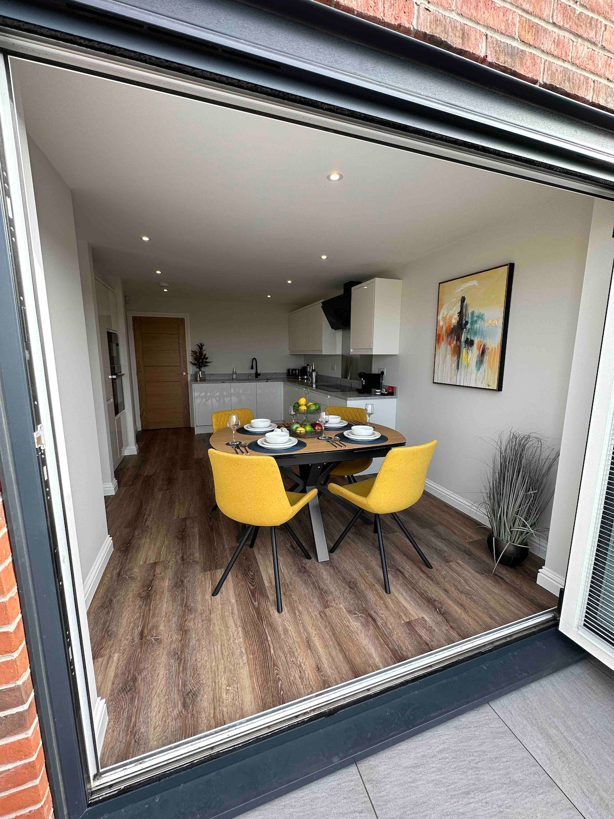 A dining area is visible through open glass doors, showcasing a round table set with dinnerware and surrounded by four yellow chairs. A modern kitchen can be seen in the background, complemented by neutral tones and artistic wall decor, creating a bright and inviting space.