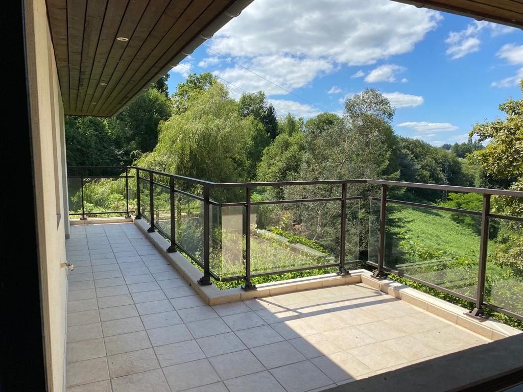 A spacious terrace features a combination of paving and railings, providing views of the surrounding greenery. Sunlight creates contrasting shadows across the floor. Lush trees and open fields are visible in the background, contributing to a serene outdoor environment.
