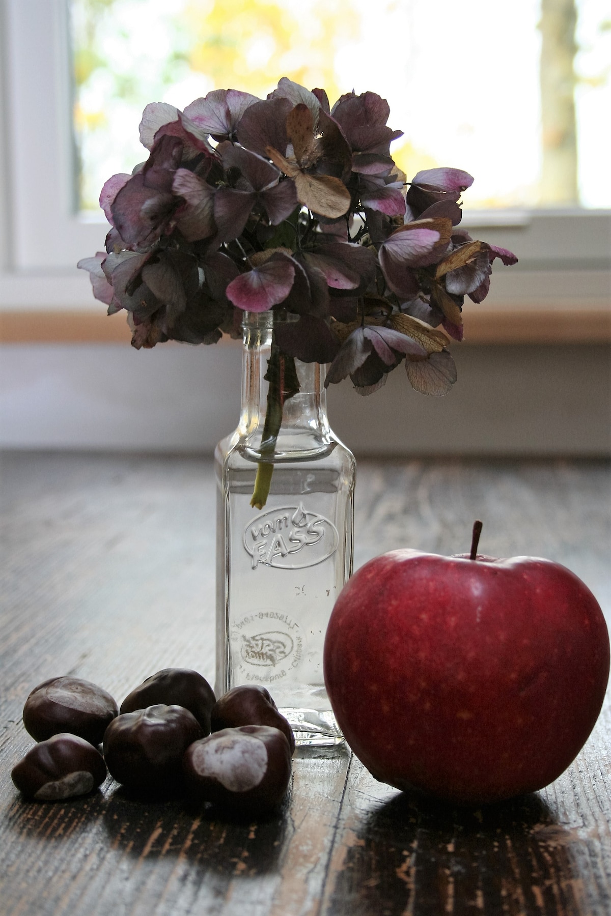 A vintage glass bottle holds dried purple flowers, set on a wooden surface. Next to the bottle, a red apple rests alongside several chestnuts, creating a calm and natural still life composition.