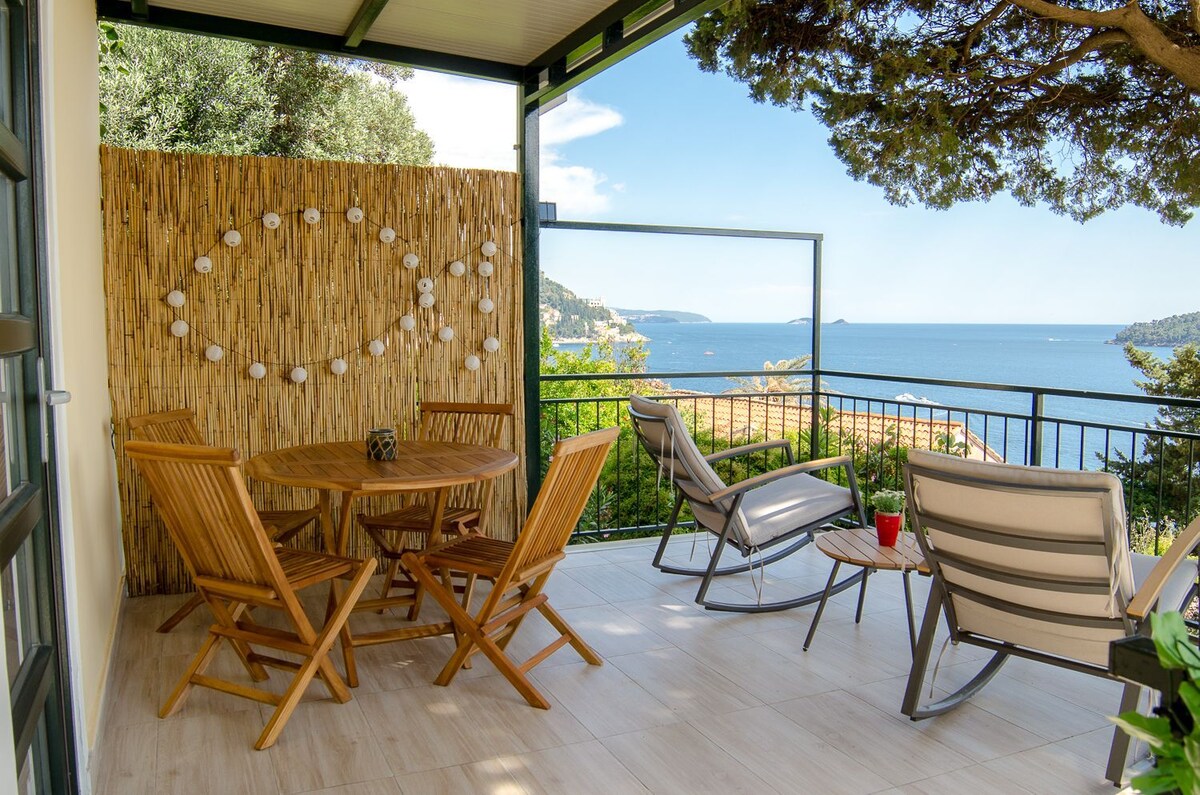A private terrace is displayed, featuring wooden patio furniture including a round table and several comfortable chairs. The railing offers unobstructed views of the turquoise sea and distant islands, while a bamboo wall adds a touch of nature. The space is bright and inviting.