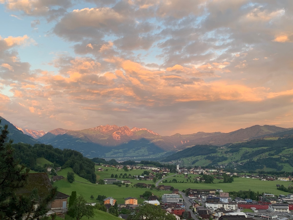 A scenic view of rolling hills and valleys is captured at sunset, showcasing soft clouds and vibrant colors in the sky. The distant mountains are illuminated by the setting sun, while the outline of the village is visible below, presenting a peaceful landscape.