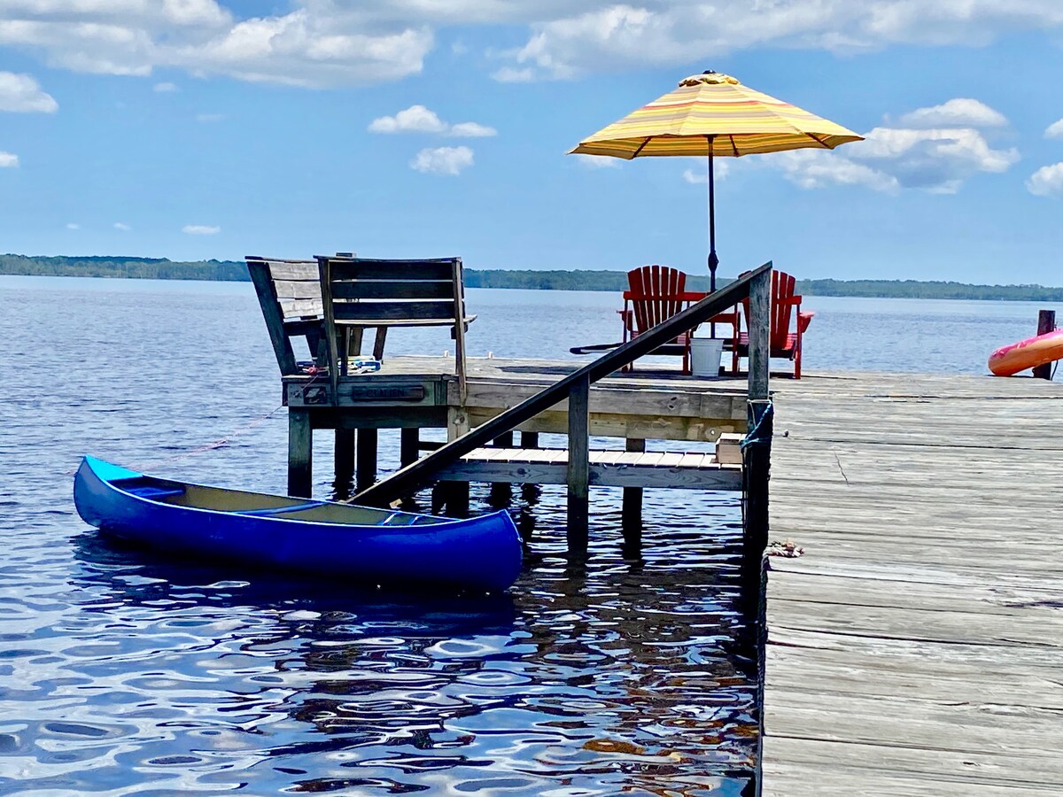 A wooden pier extends over the water, featuring a blue kayak moored alongside. Two vibrant red adirondack chairs are sheltered under a yellow striped umbrella, offering a relaxing spot to enjoy the river views on a sunny day.