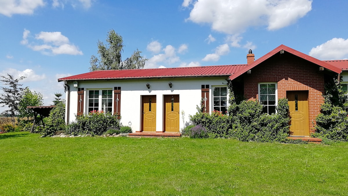 A charming building is set against a clear blue sky, featuring a red roof and cream-colored walls. Two welcoming entrance doors are framed by windows, surrounded by lush greenery and colorful flower beds. A spacious lawn invites relaxation and outdoor activities.