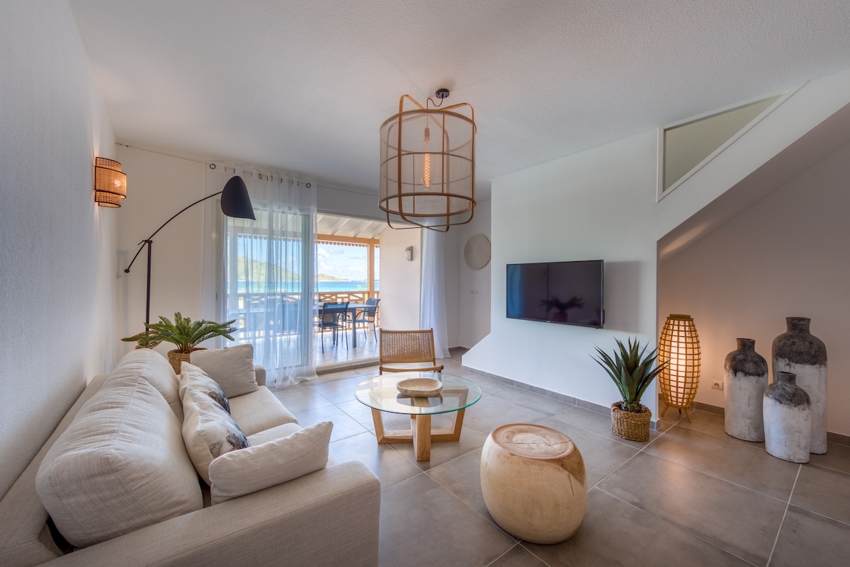A modern living space is showcased with a spacious sofa, a round glass coffee table, and decorative plants. Large windows allow natural light to fill the area. An entertainment unit is mounted on the wall, enhancing the cozy atmosphere.
