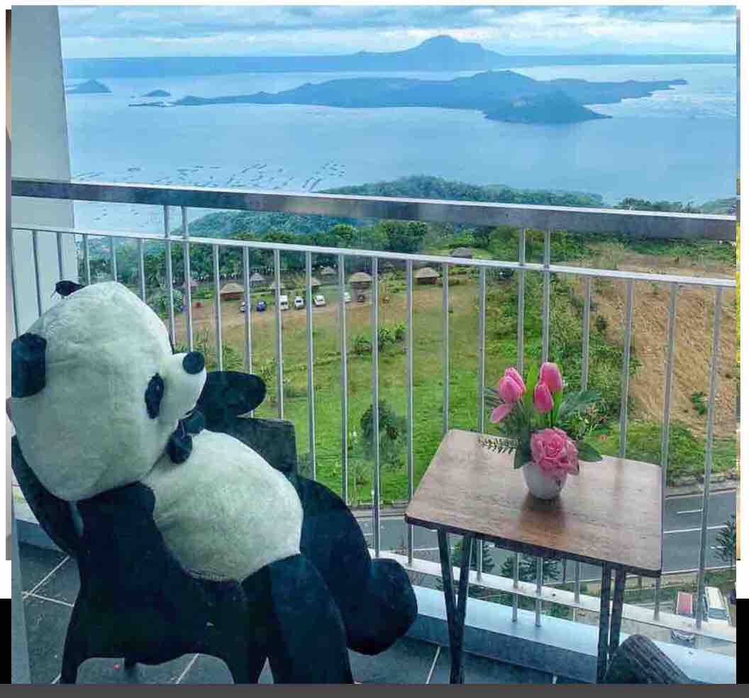 A comfortable balcony is presented, featuring a large panda-shaped chair overlooking a serene view of Taal Volcano and the surrounding landscape. A small wooden table holds a vase with fresh pink flowers, creating a peaceful outdoor setting.