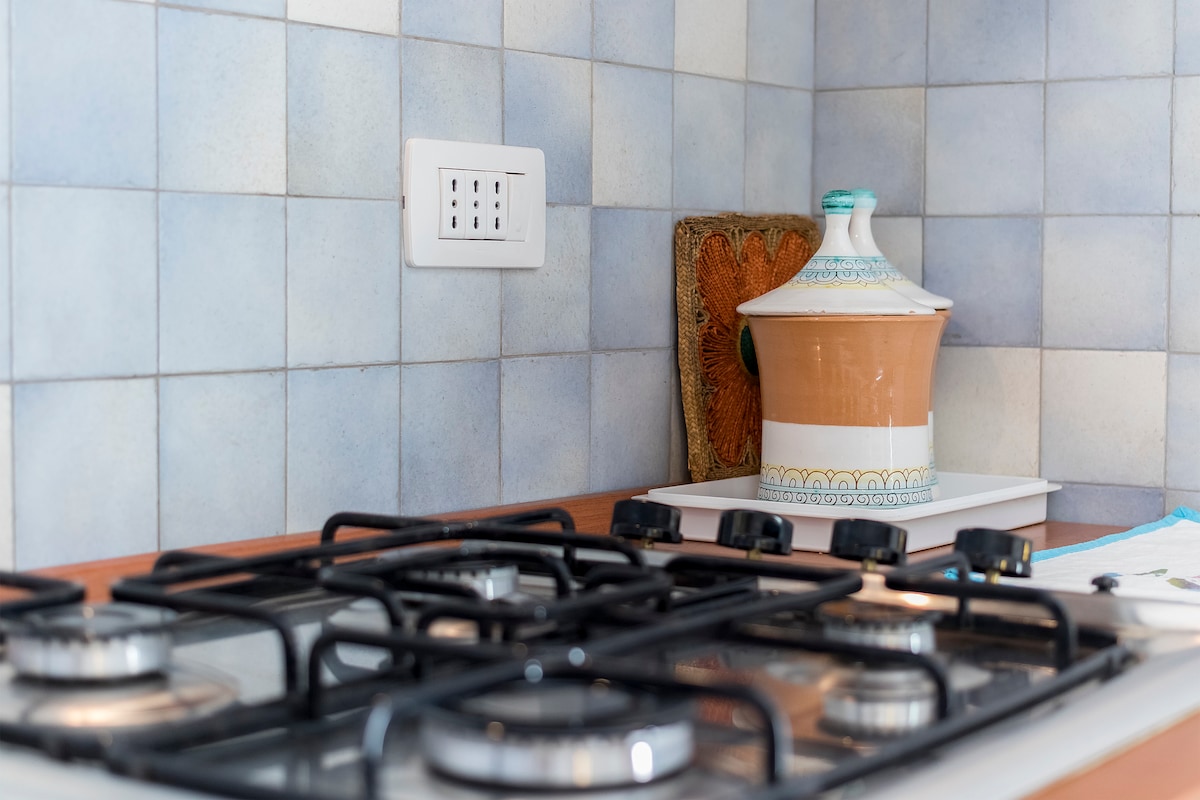 A well-equipped kitchen features a gas stove with multiple burners and a traditional ceramic container in soft tones. Light blue tiles cover the walls, providing a serene backdrop. A power outlet is conveniently located nearby, enhancing functionality.