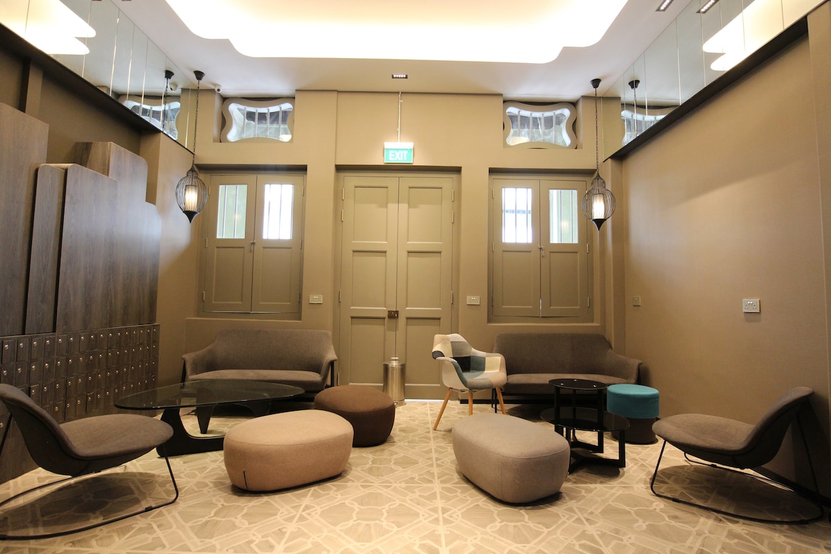 A communal lounge area features a variety of seating options, including curved chairs and soft ottomans. The light-colored walls and mirrored accents enhance the space's openness. Soft ambient lighting is provided by lantern-style fixtures, creating a comfortable environment for guests to relax and socialize.