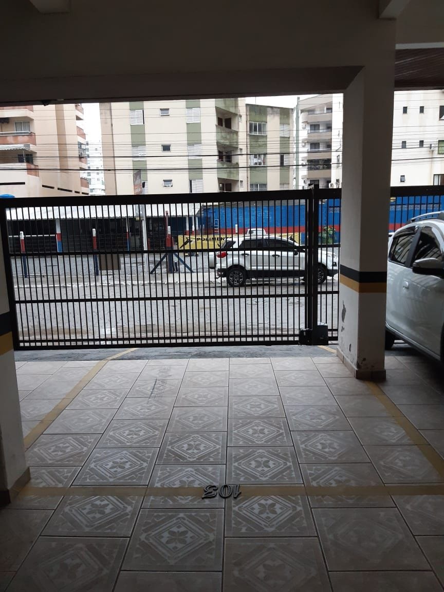 A covered parking area is visible, featuring tiled flooring and markings indicating designated parking spots. The gated entrance allows access to the outside, where a street and buildings can be seen in the background. A parked vehicle is positioned within the parking space.