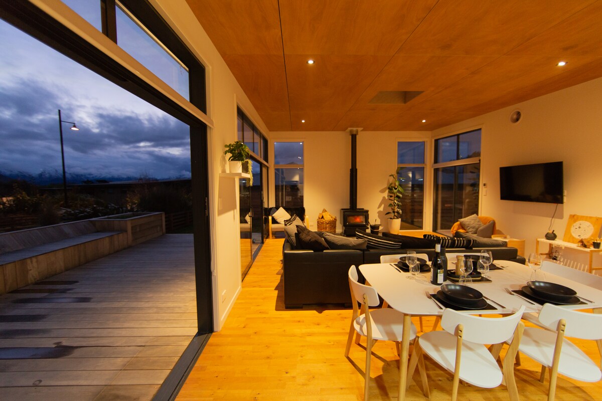 A spacious living and dining area features large windows with views of the outdoor patio. A cozy seating arrangement surrounds a fireplace, while a dining table is set with plates and cutlery. The wood ceiling and warm wooden floor create a welcoming atmosphere.