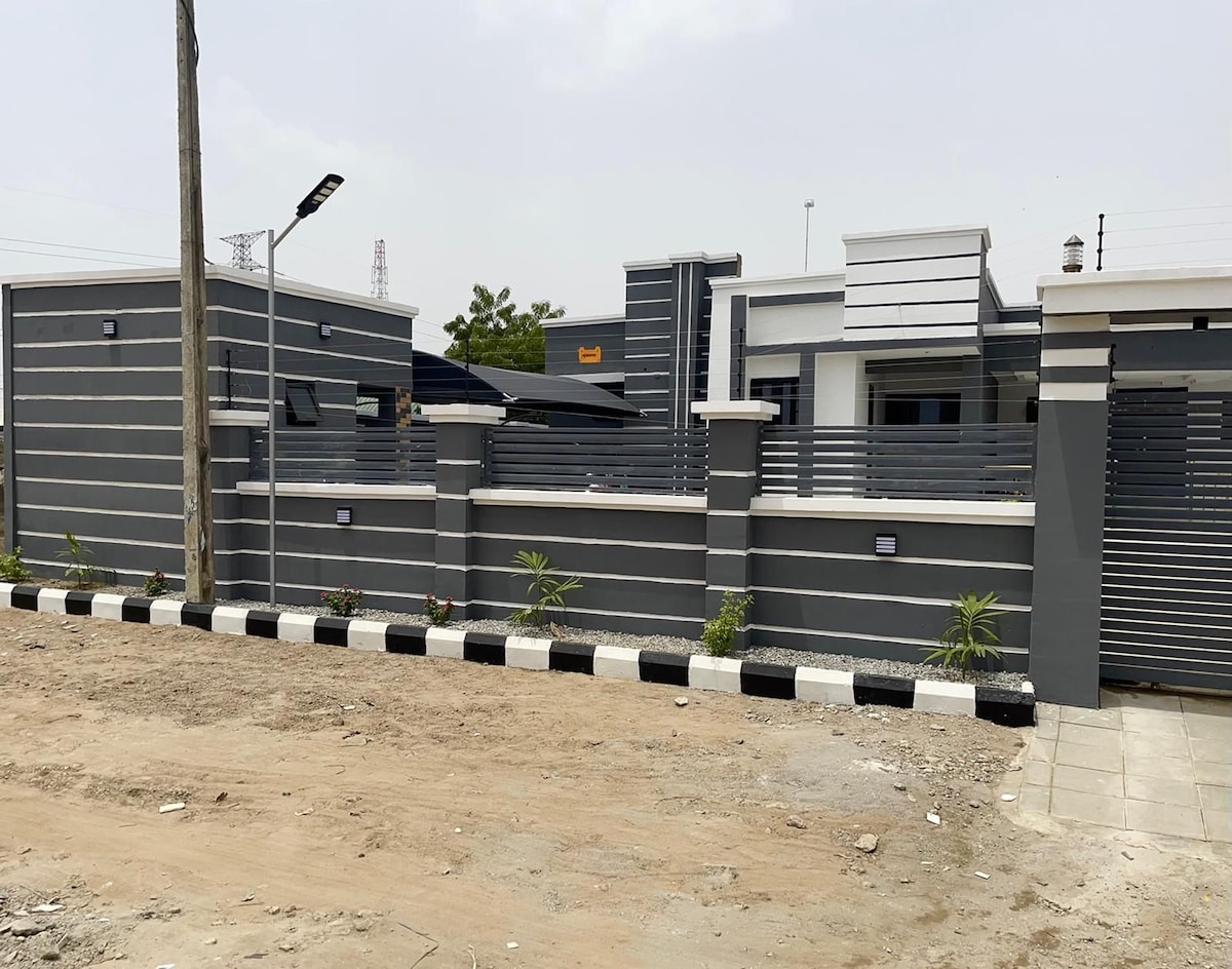 A modern exterior showcases a gray facade with horizontal stripes. The property is enclosed by a low fence, accented by small plants along the pathway. A light pole is positioned on the left, while the entrance gate features a contemporary design.
