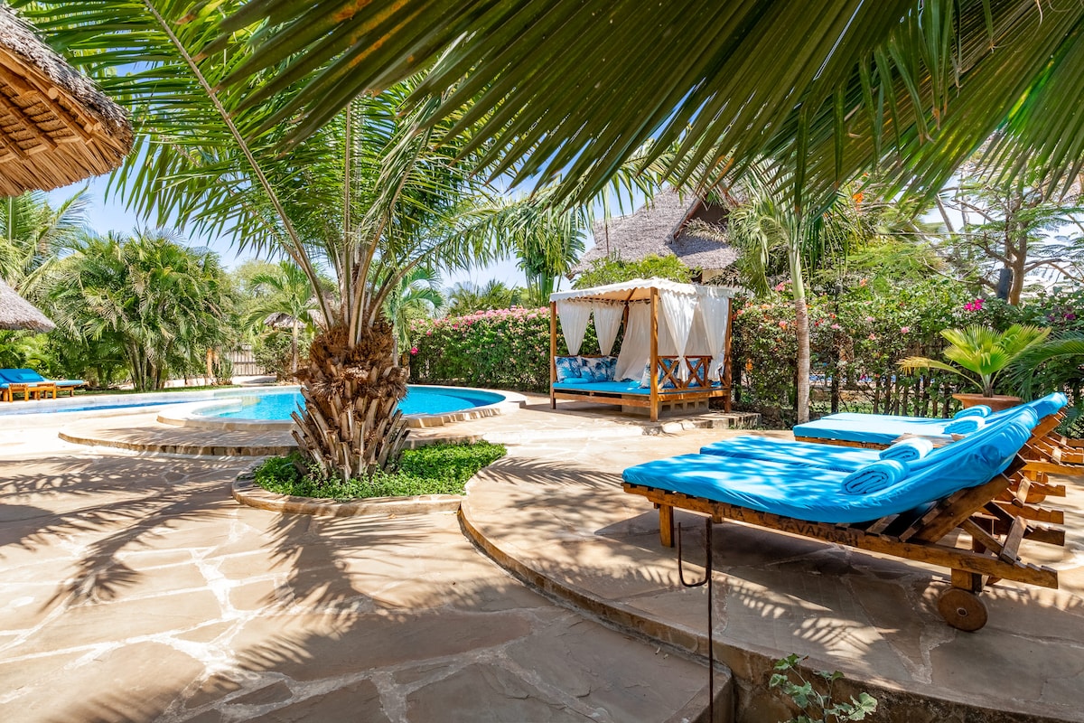 A private pool area is framed by lush greenery and tropical palms. Comfortable loungers with bright blue cushions are positioned around the pool, providing ample space for relaxation. A shaded cabana is visible in the background, enhancing the serene outdoor setting.