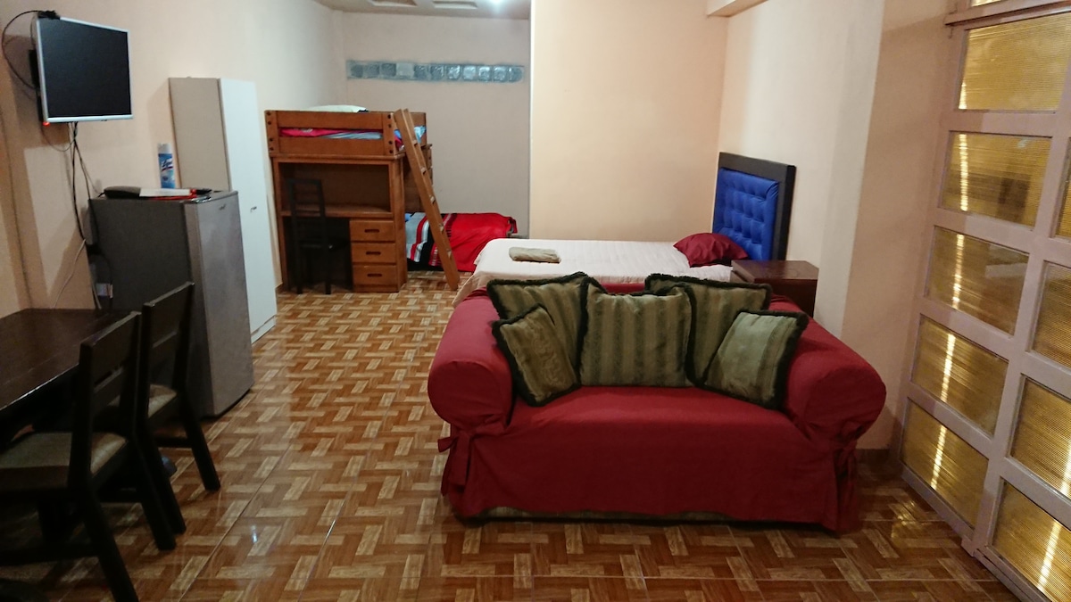 The image showcases an open-layout living space featuring a double bed alongside a bunk bed with individual mattresses. A red couch with decorative pillows occupies the center, complemented by a small dining area with a table and four chairs. A television is mounted on the wall.