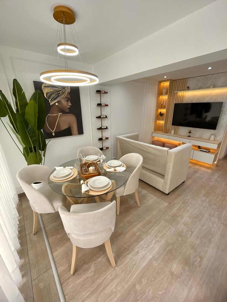 A dining area features a glass table surrounded by four light-colored chairs, set with dinnerware. A decorative plant adds a touch of greenery. A cozy living area is visible in the background, showcasing a stylish sofa and a media unit with soft lighting.