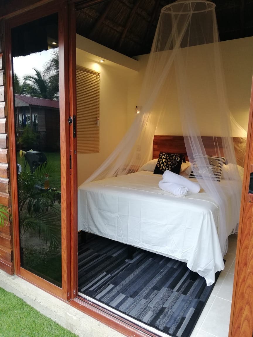 A bright sleeping area is enclosed with a mosquito net, showcasing a spacious bed dressed in white linens. Natural light enters through large glass doors, opening into a serene outdoor view with tropical greenery.