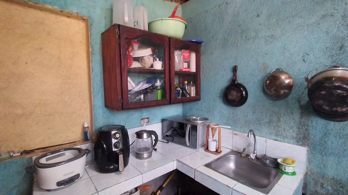 A compact kitchen features wooden cabinets displaying various dishes and kitchen appliances. Counter space includes a microwave, coffee maker, and a sink. Metal pots hang on the wall, while a bowl of fresh fruit is placed on the counter, contributing to a functional cooking area.