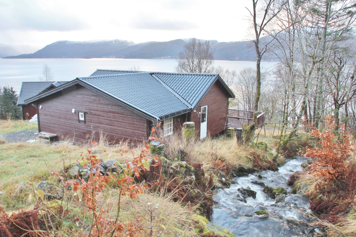 A wooden cabin with a dark roof is positioned on a hillside overlooking a calm body of water. Nearby, a gentle stream flows past, surrounded by natural foliage and rocky terrain. The backdrop features distant mountains and a cloudy sky.