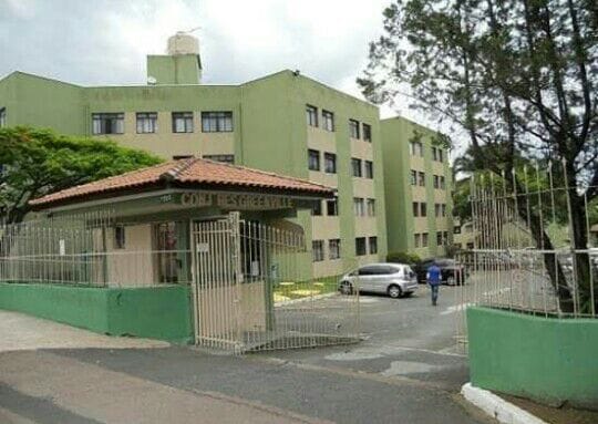 A multistory building with a green exterior is shown from the entrance, featuring a gated entryway. A tiled roof structure is visible at the front, bordered by trees and shrubbery. A pathway leads into the property, where a person walks towards the building.