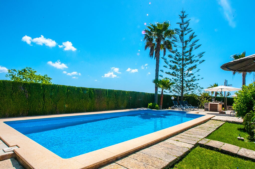 A spacious outdoor pool is framed by lush greenery and palm trees, inviting relaxation under the clear blue sky. Sun loungers are arranged along the edge, while a shaded seating area is visible nearby, providing a tranquil space for rest and leisure.
