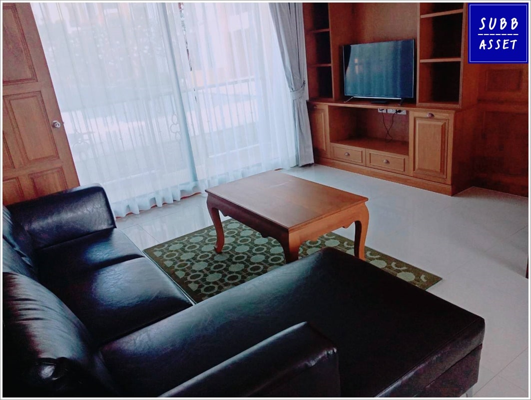 A living area features a dark leather sofa and a wooden coffee table set on an area rug. Natural light filters through sheer curtains covering large windows, with a television mounted on a wooden media unit visible in the background.