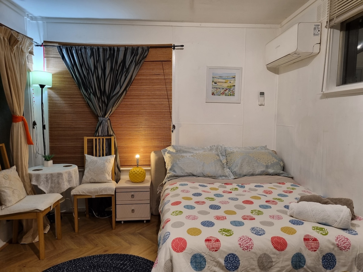 A cozy sleeping area is presented, featuring a spacious bed with a multicolored blanket. Two chairs and a small table are positioned beside the bed. A lamp provides soft lighting, while patterned curtains frame a window. A wall-mounted air conditioner is visible.