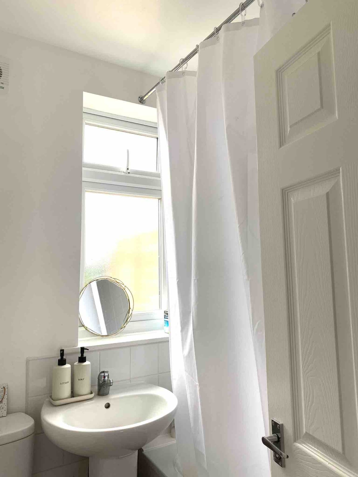A bathroom setting is displayed, featuring a clean white aesthetic. A circular mirror hangs above a small sink, complemented by neatly arranged toiletries. Natural light enters through a window, partially obscured by a white shower curtain, creating an airy and bright environment.