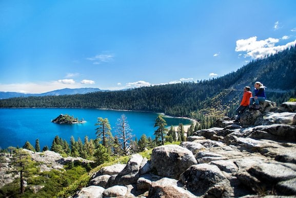 A scenic view of Lake Tahoe is captured from a rocky outcrop. Two individuals are seen sitting near the edge, overlooking the sparkling blue water and a small island surrounded by tall trees. The mountains provide a serene backdrop under a clear blue sky.