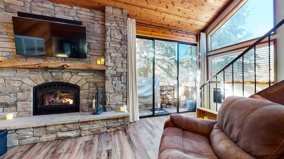 A stone fireplace is positioned beneath a wall-mounted television in a warm living space. Large glass doors offer natural light and a view of the exterior. Comfortable seating includes a plush sofa, and the wood-paneled ceiling enhances the room's rustic appeal.