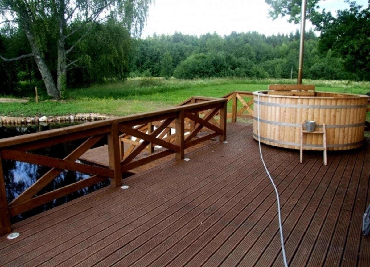 A wooden deck extends over the water, featuring a circular wooden hot tub positioned beside the edge. The area is bordered by wooden railings, while lush greenery and trees are visible in the background, enhancing the natural setting.