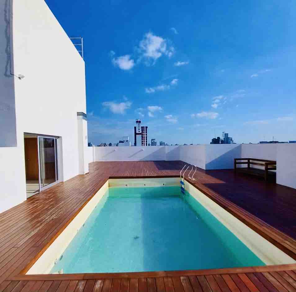 A rooftop terrace features a clear swimming pool surrounded by warm wooden decking. The sky is bright and expansive, with scattered clouds adding texture. Nearby, a simple bench offers a space to sit and enjoy the view of the city skyline.