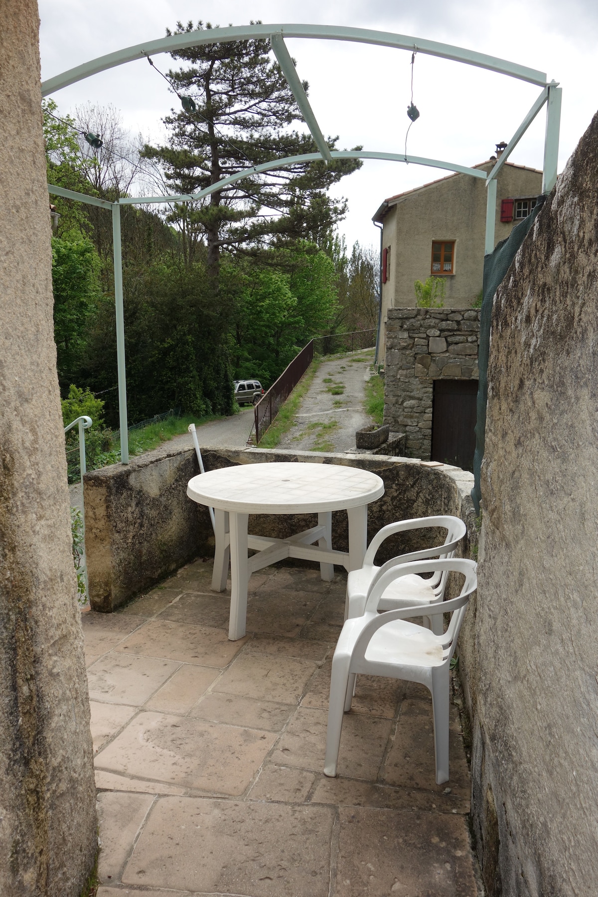 An outdoor terrace features a round table surrounded by three white chairs. The area is bordered by stone walls, and greenery is visible in the background. A quiet road is seen in the distance, contributing to a serene setting.