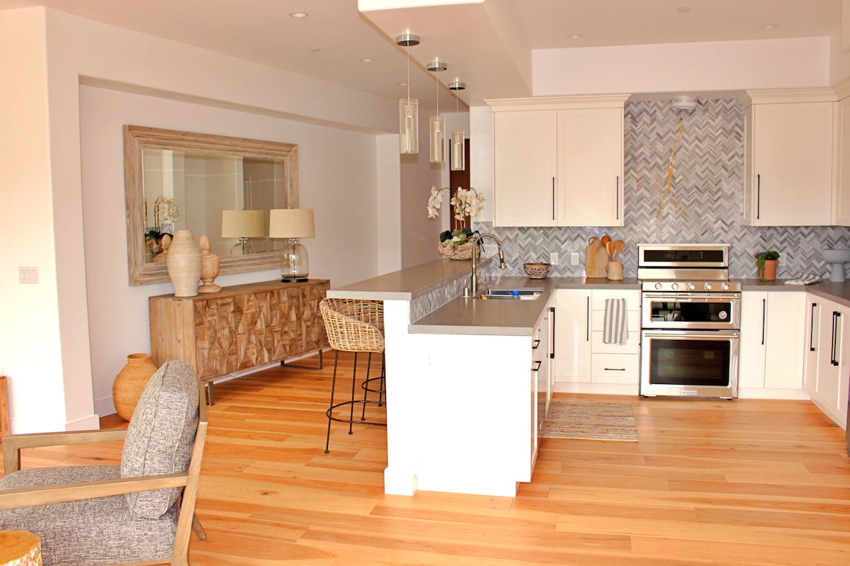 A modern kitchen is featured with sleek cabinetry, state-of-the-art appliances, and a spacious island with seating. Natural wood flooring adds warmth, while a large mirror reflects light across the space. Decorative items are placed on a side console, enhancing the contemporary design.