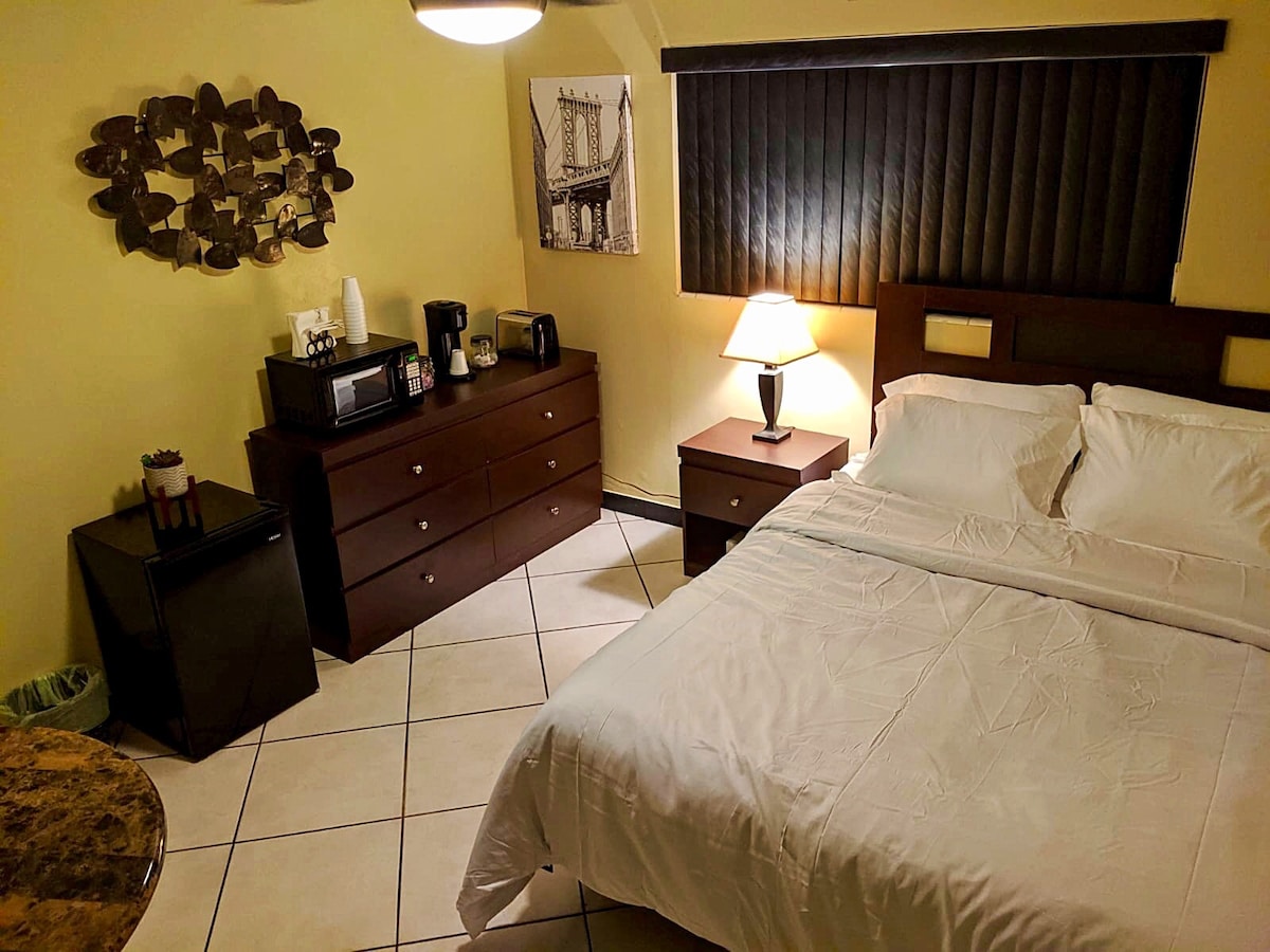 A well-arranged master bedroom features a spacious bed accompanied by bedside tables. A dresser stands against the wall, with a microwave and coffee station positioned nearby. Soft lighting is provided by a table lamp, and a collage of circular wall decor adds visual interest.