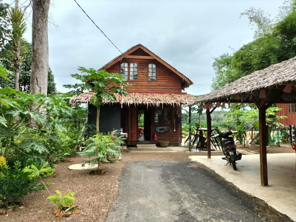 A rustic wooden building with a thatched roof is set amidst greenery, featuring a welcoming entrance. A stone pathway leads to the door, flanked by various plants and trees. A covered area for parking and outdoor relaxation is visible to the right.