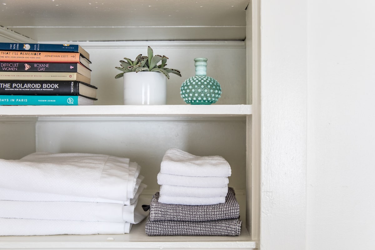 A white shelf displays neatly arranged white towels and a set of folded patterned dish towels. A small potted plant and a decorative glass piece add a touch of color. Multiple books on various subjects are stacked upright, enhancing the organized look.