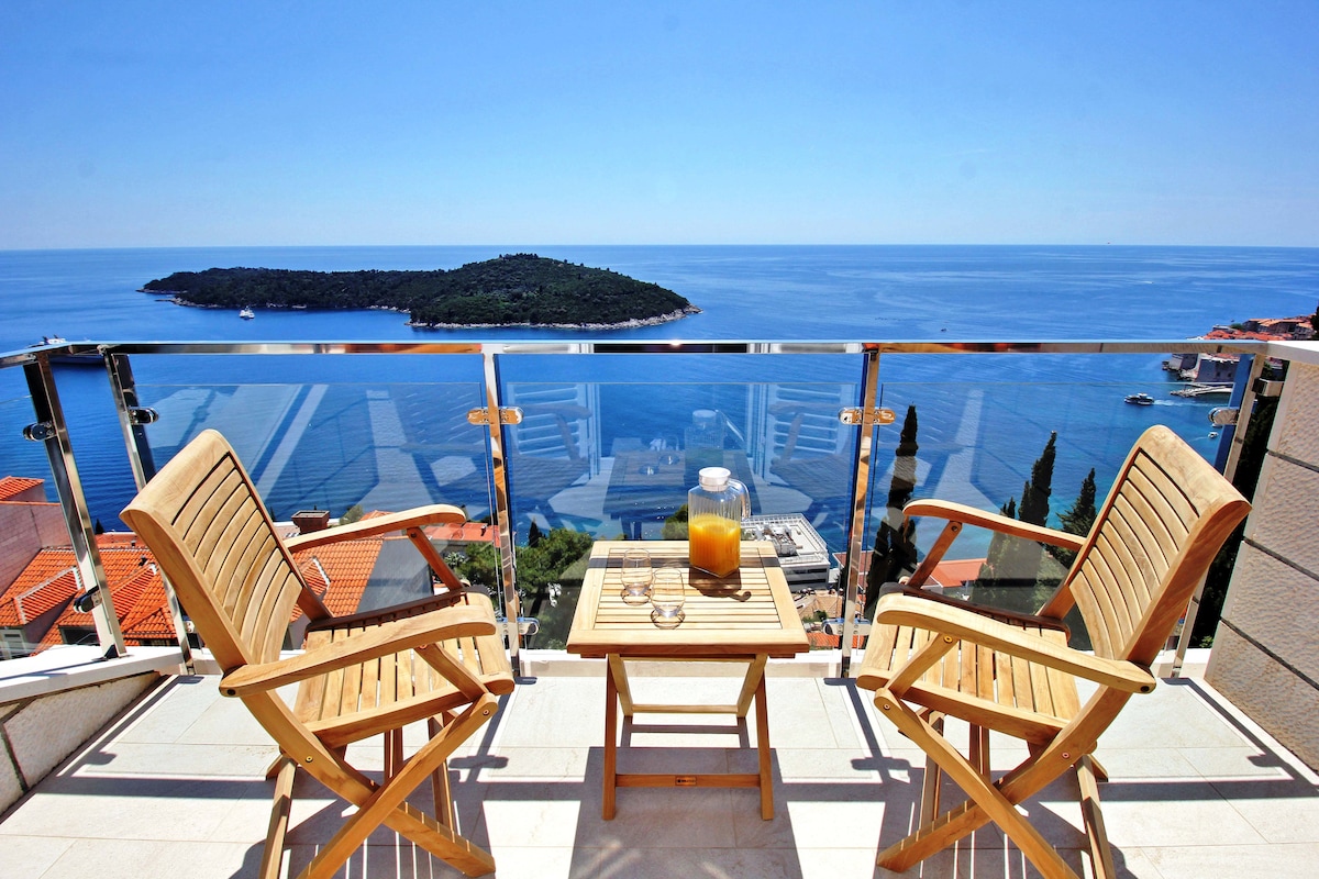 A balcony is shown with two wooden armchairs and a small table. A pitcher and glasses are placed on the table, with panoramic views of the Mediterranean and a nearby island visible across the water. The clear sky enhances the serene atmosphere.