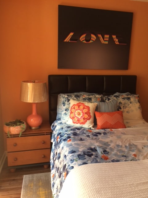 A warm bedroom is presented with an orange wall, featuring a modern art piece that spells 'LOVE.' A spacious bed is adorned with a floral comforter and decorative pillows. A nightstand is positioned beside the bed, topped with a lamp and a potted plant.