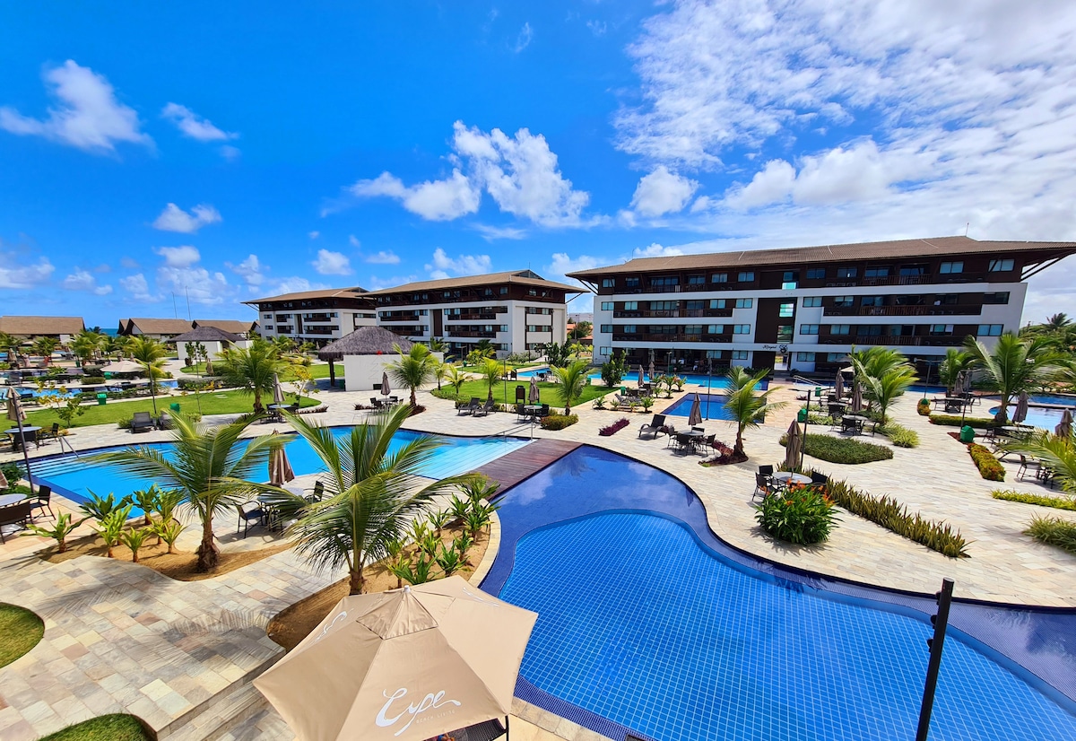 The resort features a spacious outdoor area with multiple swimming pools surrounded by lush greenery and palm trees. Lounge chairs are positioned around the pools, offering a relaxing atmosphere. Modern buildings are visible in the background under a clear blue sky.