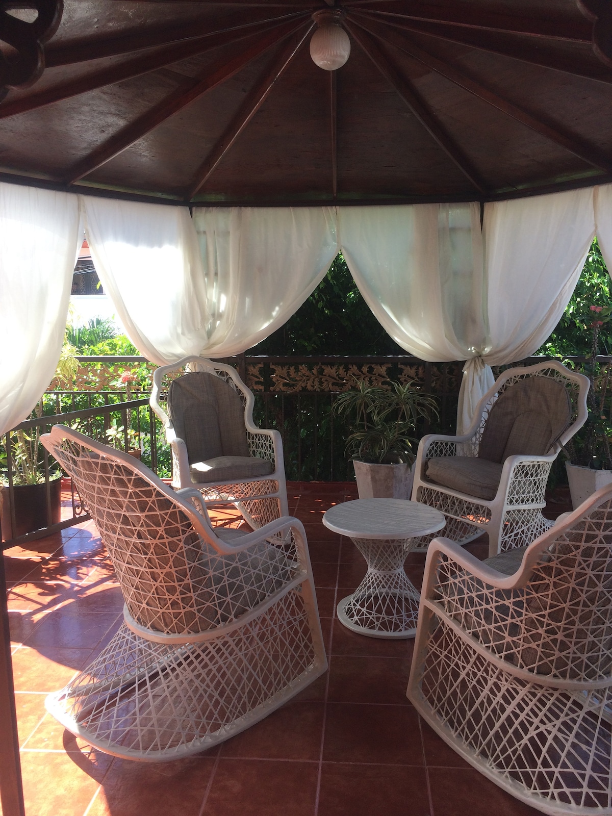 A covered seating area features four woven chairs arranged around a circular table. Sheer white curtains create a soft backdrop, with greenery visible beyond the railing, adding a natural element to the space.