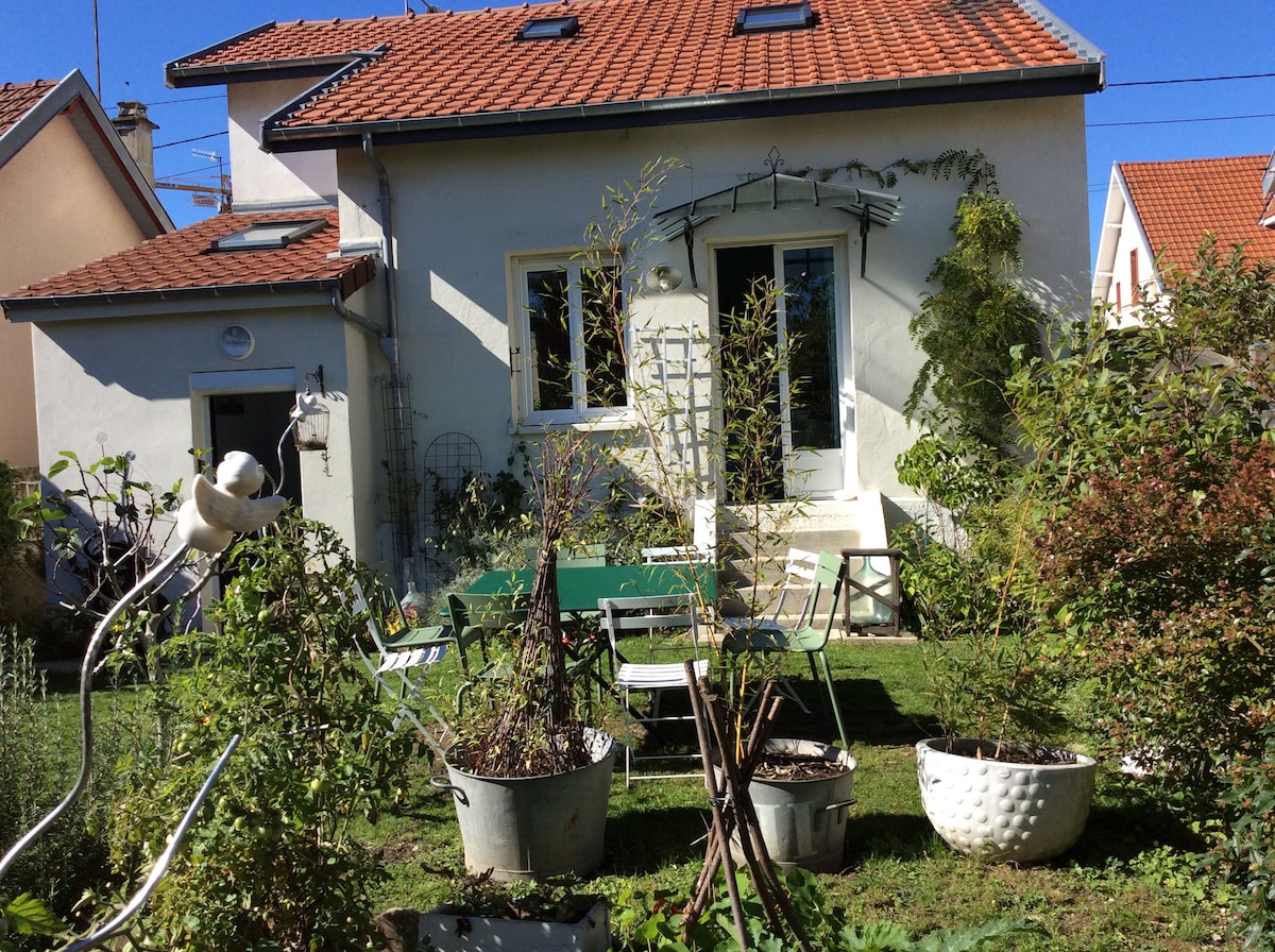 A charming house is visible with a red tiled roof, surrounded by a landscaped garden. Various plants and trees are arranged in planters, alongside outdoor seating. Large glass doors open to a green outdoor space, inviting natural light into the interior.