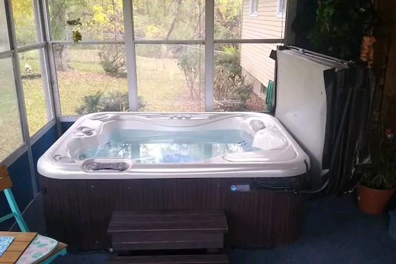 A hot tub is situated in a screened-in area, surrounded by greenery visible through the screens. The tub features a spacious design with steps leading up to it. Natural light filters in, creating a calming atmosphere. Various plants are placed nearby.