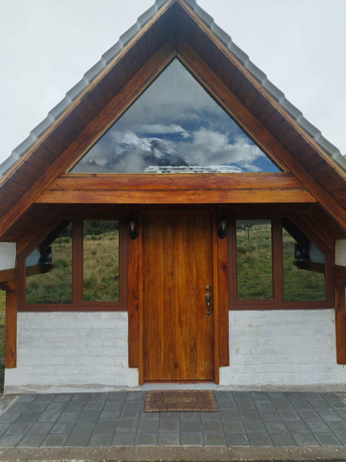The distinctive cabin entrance features a triangular wooden structure with a glass-panel peak, reflecting the cloudy sky above. Two lantern-style lights are mounted alongside the door, which is framed by large windows, offering views of the surrounding natural landscape.