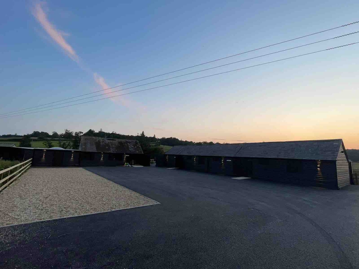 The image showcases a row of newly renovated agricultural barns set against a pastel sky at sunset. A spacious gravel area is visible in the foreground, providing access to the barns, which exhibit dark timber exteriors and are situated within a serene countryside landscape.