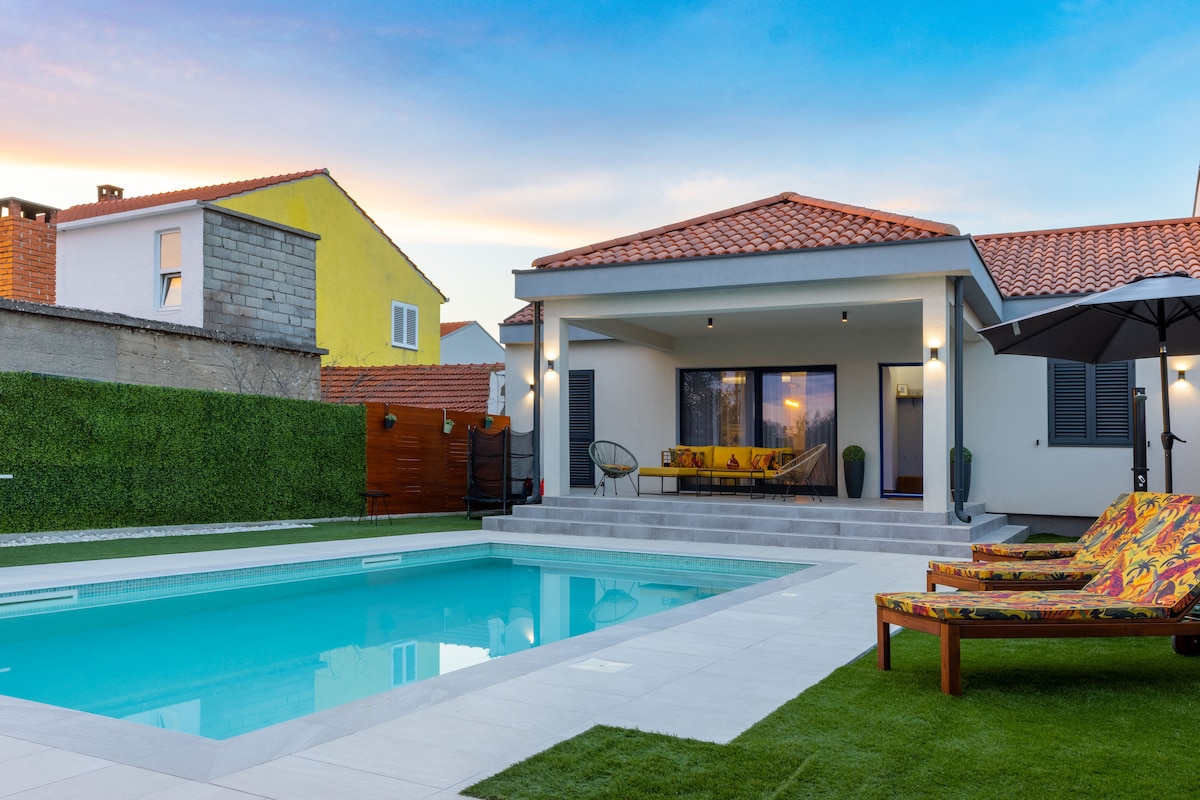 A modern villa is shown with a pristine pool reflecting the evening sky. Comfortable lounge chairs in vibrant colors are positioned beside the pool, surrounded by manicured grass. The exterior features a spacious patio with large glass doors that open into the indoors.