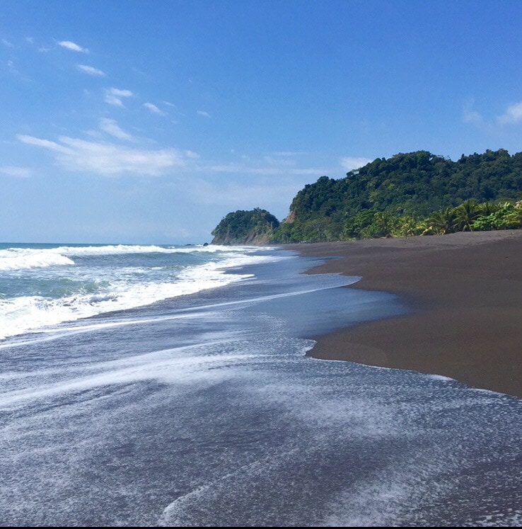A serene beachfront scene is captured, featuring gentle waves lapping at the shore of a dark sand beach. Lush green hills rise in the background, contrasting with the clear blue sky and soft white clouds.