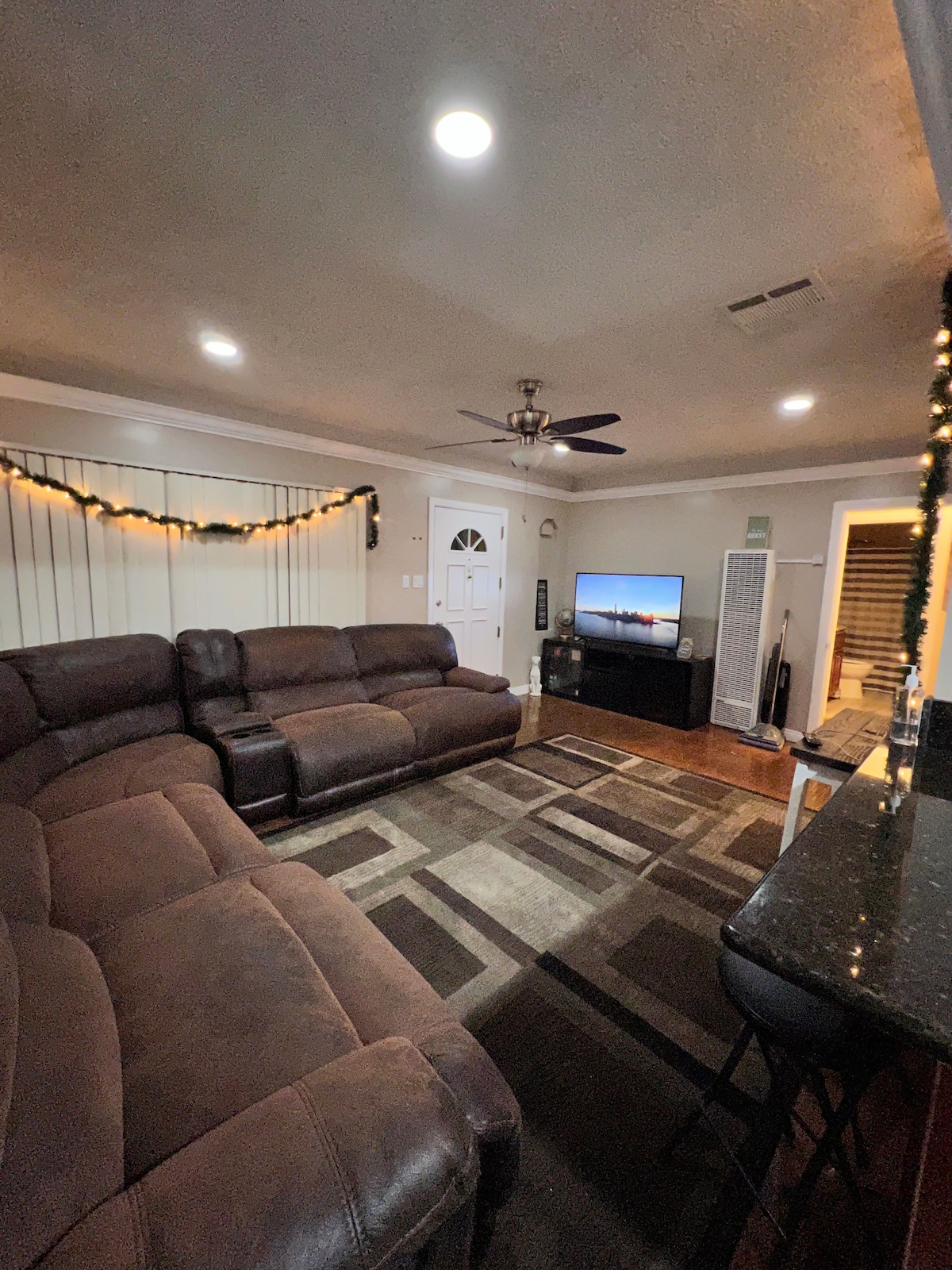 A spacious living room features a large sectional couch, with decorative lighting draped along the walls. A plasma TV is positioned on a stand, while a patterned area rug adds comfort underfoot. Natural light is allowed in through blinds on the windows.