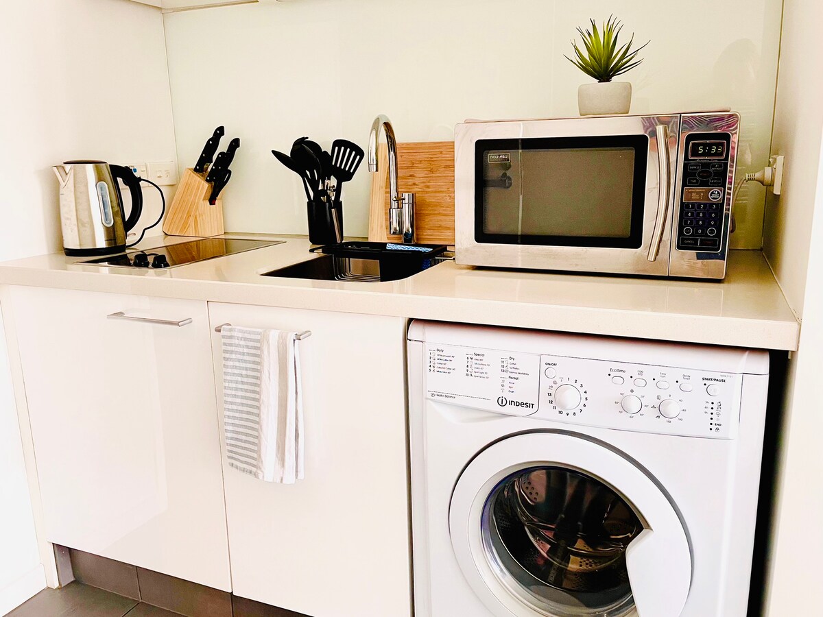 A functional kitchenette is displayed, featuring a built-in sink, a microwave, and an electric kettle. Cooking utensils are organized in a holder. A washing machine is situated beneath the counter, marked by a clean and modern design. A small potted plant adds a touch of greenery.